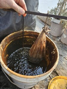 A picture of a five gallon bucket filled partially with compost tea. A hand is holding a stick with a bag of compost hanging from it.